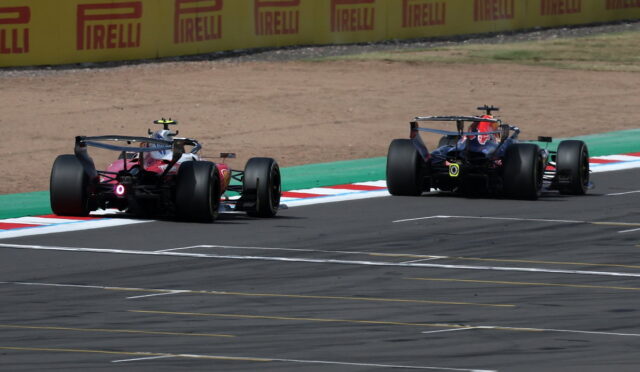 Two Formula 1 cars race side by side on a track, with yellow Pirelli barriers in the background.