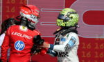 Ferrari driver Charles Leclerc in a red suit shakes hands with a rival in a white racing suit and neon helmet on a red backdrop at the podium.