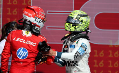 Ferrari driver Charles Leclerc in a red suit shakes hands with a rival in a white racing suit and neon helmet on a red backdrop at the podium.