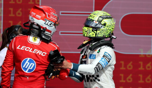 Ferrari driver Charles Leclerc in a red suit shakes hands with a rival in a white racing suit and neon helmet on a red backdrop at the podium.