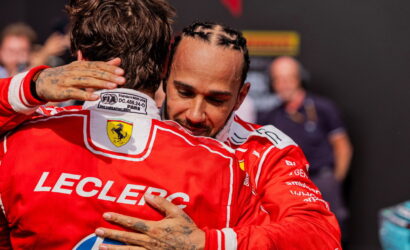 Two Formula 1 drivers in red Ferrari racing suits hug each other, their faces close in a moment of celebration, with one jacket showing a crest and the word 'LECLERC' on the back.