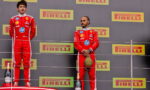 Two Formula 1 drivers stand on a podium in red Ferrari suits, with champagne bottles at their feet and Pirelli banners in the background.
