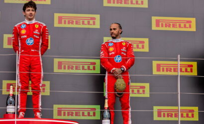 Two Formula 1 drivers stand on a podium in red Ferrari suits, with champagne bottles at their feet and Pirelli banners in the background.