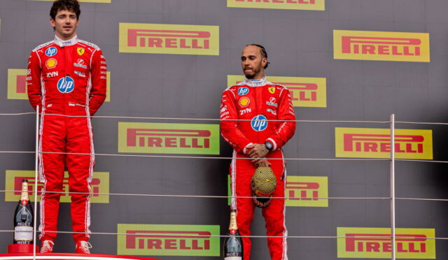 Two Formula 1 drivers stand on a podium in red Ferrari suits, with champagne bottles at their feet and Pirelli banners in the background.