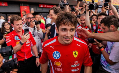 Ferrari driver in a red team shirt surrounded by reporters and photographers taking pictures.