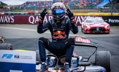 Race driver in Red Bull suit sits on a go-kart at the track, hands raised in victory with cheering grandstands behind them in the pit area.