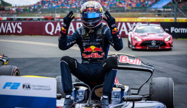 Race driver in Red Bull suit sits on a go-kart at the track, hands raised in victory with cheering grandstands behind them in the pit area.