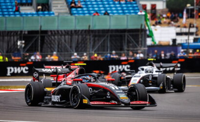 Front black and red Formula race car leads a tight bend, with two other cars close behind on a racing circuit; empty blue grandstands in the background.