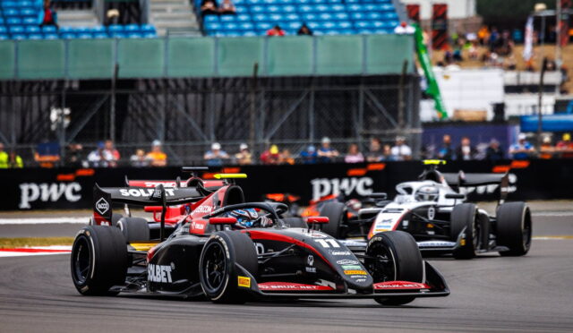 Front black and red Formula race car leads a tight bend, with two other cars close behind on a racing circuit; empty blue grandstands in the background.