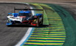 BMW race car taking a tight left-hander on a track, blue and yellow striped curbs visible on the inside.