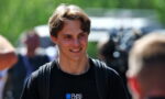 Young man with a backpack smiling outdoors at a sunny event with blurred crowd in the background, foreground in focus.
