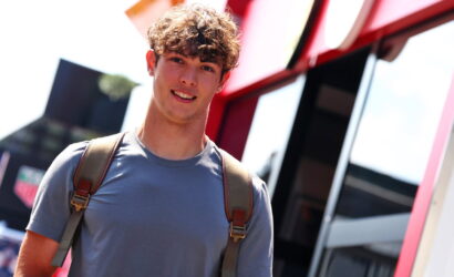 Young man with a brown backpack and gray t-shirt standing beside a red bus, smiling outdoors.