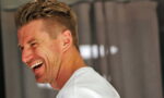 Side-profile of a man with short blonde hair laughing, wearing a white shirt, teeth visible as he smiles.