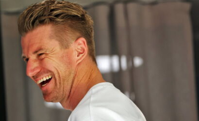 Side-profile of a man with short blonde hair laughing, wearing a white shirt, teeth visible as he smiles.