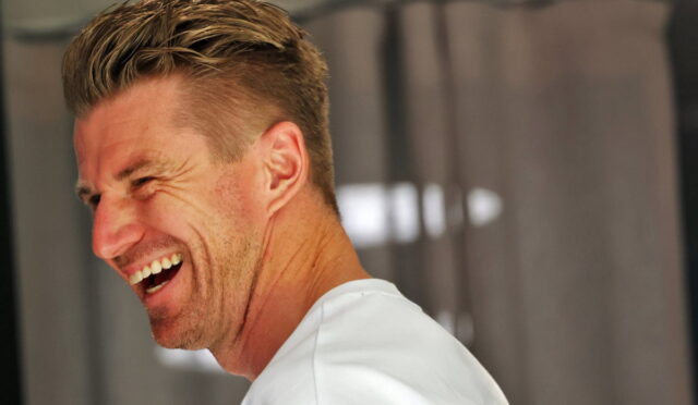 Side-profile of a man with short blonde hair laughing, wearing a white shirt, teeth visible as he smiles.