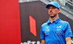 Young man in a blue racing polo and cap stands in front of a red and black backdrop at what looks like a race event.