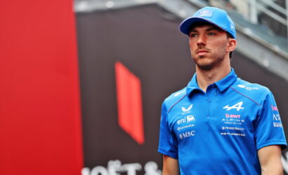 Young man in a blue racing polo and cap stands in front of a red and black backdrop at what looks like a race event.