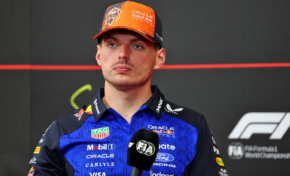 Portrait of a Formula 1 driver in a blue sponsor-laden uniform and orange cap, speaking into a microphone.