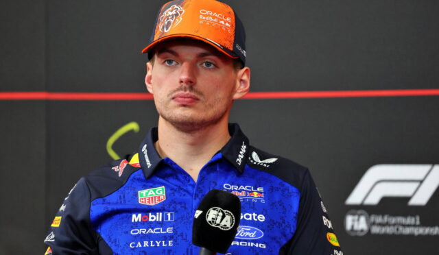 Portrait of a Formula 1 driver in a blue sponsor-laden uniform and orange cap, speaking into a microphone.
