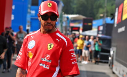 Man in red Ferrari team shirt and cap with sunglasses, standing in a paddock with blurred team tents in the background.