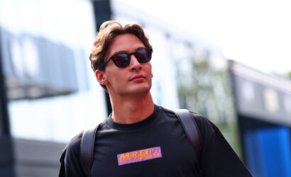 Young man in a black shirt with an orange logo, wearing sunglasses, standing in an urban, blurred street setting.