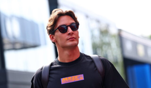Young man in a black shirt with an orange logo, wearing sunglasses, standing in an urban, blurred street setting.