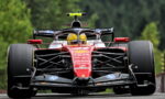 Front-on view of a red Formula 1 race car on a track, driver in a yellow helmet, large slick tires and sponsor logos visible.