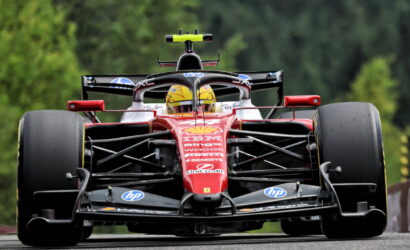 Front-on view of a red Formula 1 race car on a track, driver in a yellow helmet, large slick tires and sponsor logos visible.