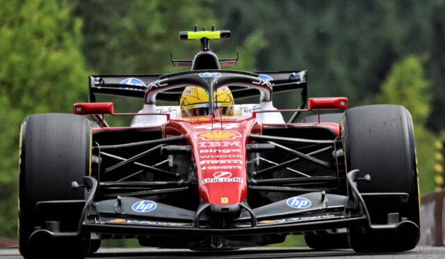 Front-on view of a red Formula 1 race car on a track, driver in a yellow helmet, large slick tires and sponsor logos visible.