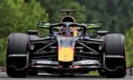 Front-on view of a Formula 1 Red Bull racing car on a track, large exposed tires and aerodynamic front wing adorned with sponsor logos (Visa), with a green, blurred forest backdrop.