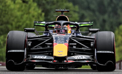 Front-on view of a Formula 1 Red Bull racing car on a track, large exposed tires and aerodynamic front wing adorned with sponsor logos (Visa), with a green, blurred forest backdrop.