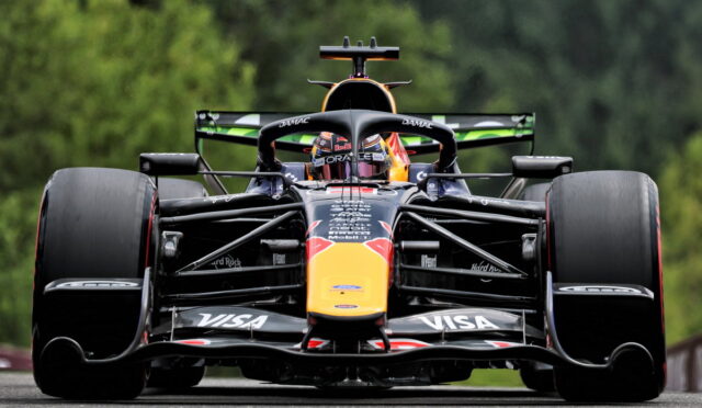Front-on view of a Formula 1 Red Bull racing car on a track, large exposed tires and aerodynamic front wing adorned with sponsor logos (Visa), with a green, blurred forest backdrop.