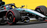 Close-up of a Mercedes Formula 1 car on track, showing the front wheel, sidepod, and aerodynamic details with a bright yellow barrier in the background