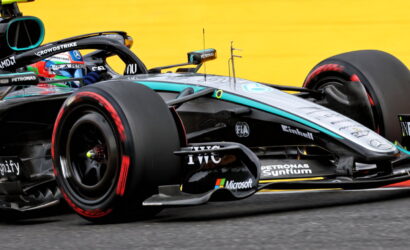 Close-up of a Mercedes Formula 1 car on track, showing the front wheel, sidepod, and aerodynamic details with a bright yellow barrier in the background