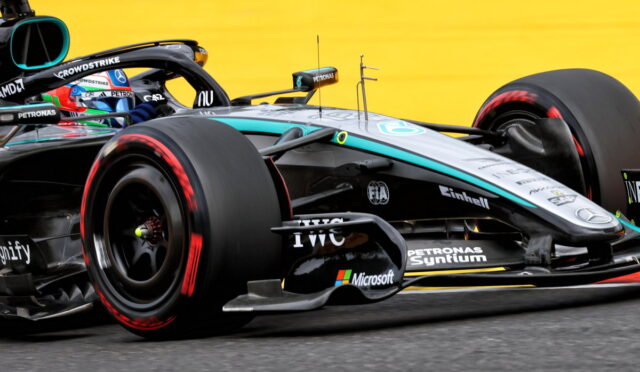 Close-up of a Mercedes Formula 1 car on track, showing the front wheel, sidepod, and aerodynamic details with a bright yellow barrier in the background