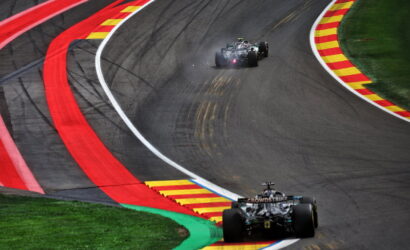Two Formula 1 cars race through a tight chicane on a track, red and yellow curb markings flashing along the bend behind a green grass runoff area behind them mandating close passing.