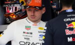 Formula 1 driver in a white suit with sponsor logos, standing in the pit area with a team member in dark Oracle Red Bull gear nearby.