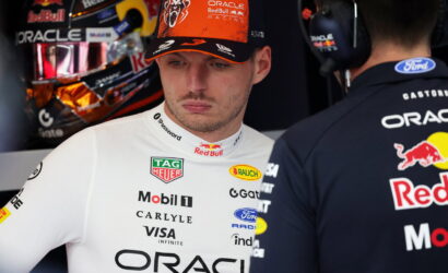 Formula 1 driver in a white suit with sponsor logos, standing in the pit area with a team member in dark Oracle Red Bull gear nearby.