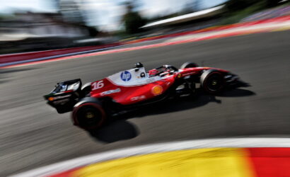 Red Formula 1-style race car speeding around a track, motion blur with red and yellow curbs in the foreground.