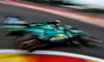 Green Formula 1 car speeding along a race track with strong motion blur, colorful barriers in the foreground and track in the background.