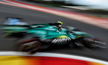 Green Formula 1 car speeding along a race track with strong motion blur, colorful barriers in the foreground and track in the background.