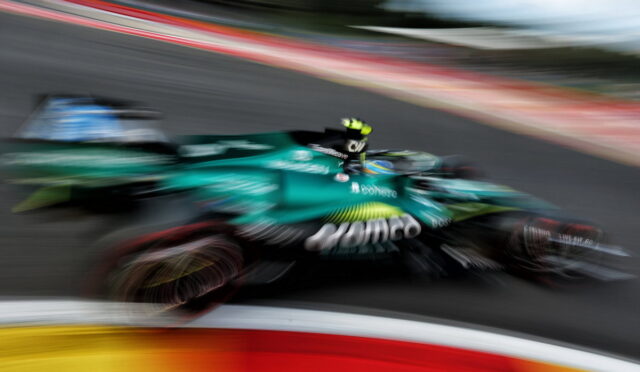 Green Formula 1 car speeding along a race track with strong motion blur, colorful barriers in the foreground and track in the background.