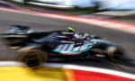 Blurred action shot of a teal and black Formula 1 car racing around a bend on a track, motion blur emphasizing speed and curvature of the circuit edge in foreground.
