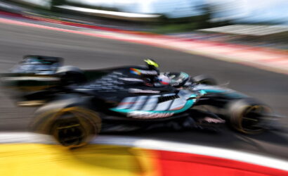 Blurred action shot of a teal and black Formula 1 car racing around a bend on a track, motion blur emphasizing speed and curvature of the circuit edge in foreground.