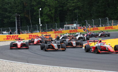Racing cars in a pack navigating a high-speed corner on a track, with DHL barriers and spectators behind a fence.