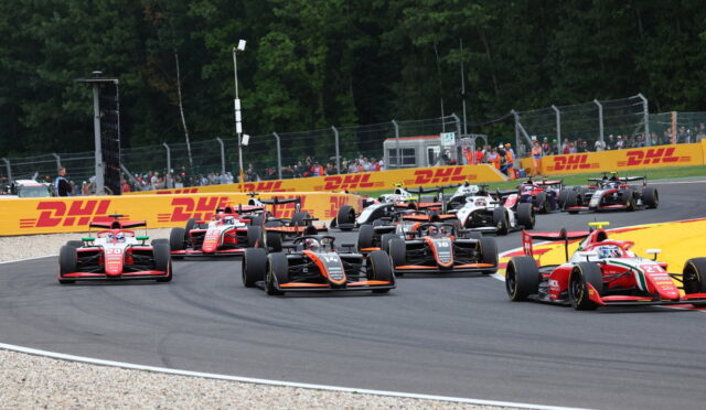 Racing cars in a pack navigating a high-speed corner on a track, with DHL barriers and spectators behind a fence.