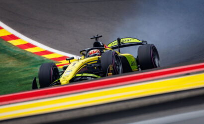 Yellow and green open-wheel race car navigates a tight corner on a race track, with red-and-yellow curbs nearby and smoke at the rear.