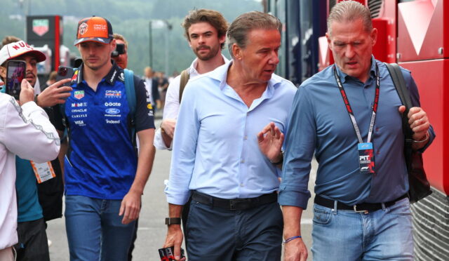 Group of men walking along a race pit lane; two in front converse while others, including a photographer, look on
