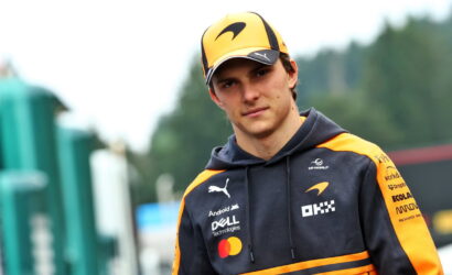 Young man wearing an orange and black racing hoodie and cap with sponsor logos, outdoors and looking at the camera.