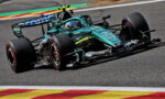 Formula 1 race car in teal and black livery speeding around a track, driver in helmet visible, on a curving corner with colorful run-off paint behind.